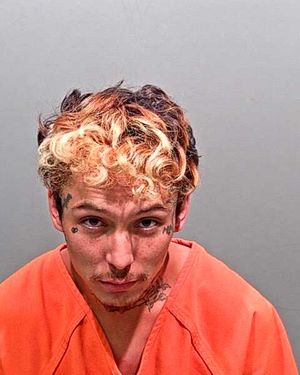 A young man with curly, dyed hair and visible tattoos is photographed in a prison orange jumpsuit against a plain background.