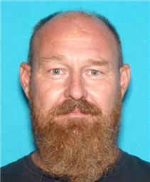 This image shows a close-up portrait of a man with a full beard and short hair, wearing a black shirt, against a light blue background.
