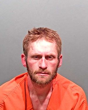 A male individual with a beard is shown in a mugshot wearing an orange jumpsuit against a gray background.