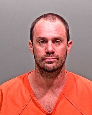 A man with short hair and a beard is wearing an orange jail uniform in a mugshot.
