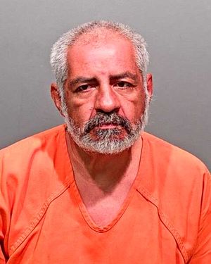 Balding man with grey bear and side hair with frown in JCSO orange jumpsuit on grey background. 
