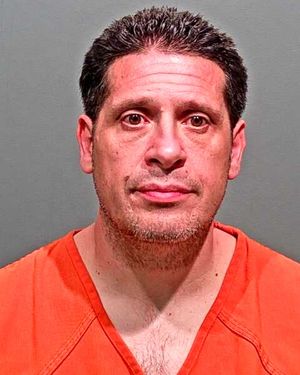A man wearing an orange incarceration jumpsuit poses for a mugshot against a plain background.