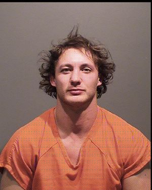 A male in an orange jail jumpsuit is posing for a mugshot against a gray background.