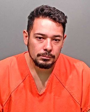 A man with short dark hair and a beard is shown wearing an orange jail jumpsuit.
