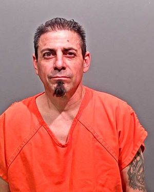 A man with dark hair, facial hair, and tattoos is wearing an orange prison jumpsuit against a gray background.