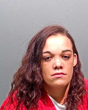 A woman with long, wavy hair is shown in a mugshot wearing a red top against a gray background.