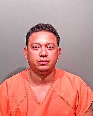 A man wearing an orange detention facility jumpsuit poses for a mugshot against a gray background.