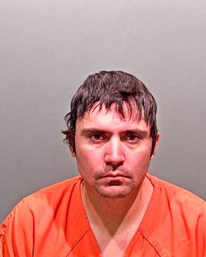 A man with short, messy hair is wearing an orange prison jumpsuit and looking directly at the camera against a gray background.
