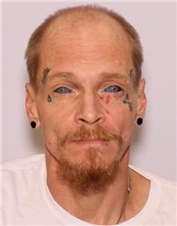 A man with facial tattoos and blue eyes, wearing a white shirt, is looking directly at the camera.