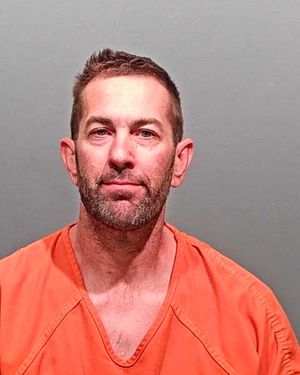 A man wearing an orange jumpsuit looks directly at the camera against a plain gray background.