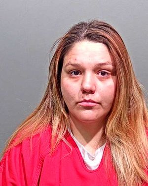 A woman with long hair, wearing a red garment, is looking directly at the camera against a plain gray background.