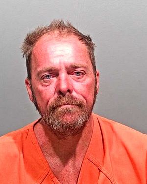 A disheveled man with a beard is shown in a mugshot, wearing an orange prison jumpsuit, against a plain background.