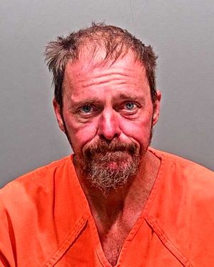 A man with disheveled hair and a beard is shown in a jail uniform, looking somber against a neutral background.