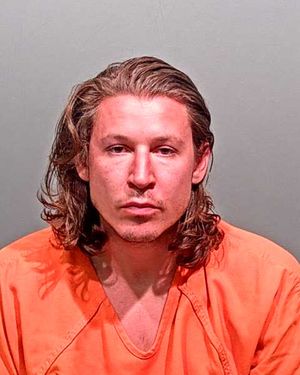 A man with long hair is posing for a mugshot while wearing an orange prison jumpsuit.