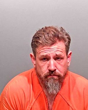 A man with a beard is shown in an orange prison jumpsuit, looking somber in a mugshot setting.