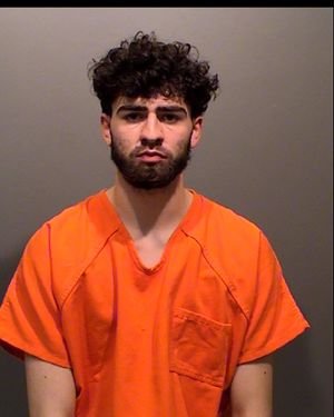 A young man with curly hair is wearing an orange jail jumpsuit and has a neutral expression.