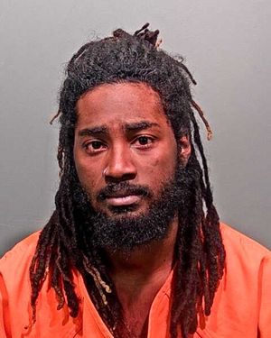 A man with long, dark hair and a beard is shown wearing an orange jumpsuit in a mugshot.