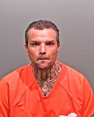 A man with tattoos on his neck and a serious expression is wearing an orange jumpsuit against a plain background.