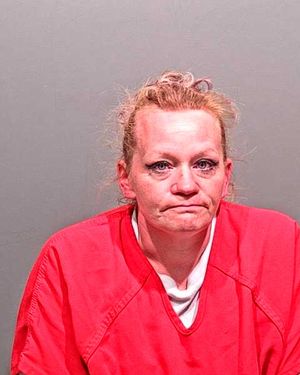 A woman with disheveled hair and a serious expression is wearing a red inmate jumpsuit against a gray background.