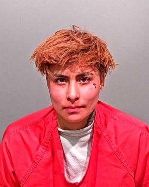 A person with light brown hair and facial tattoos is wearing a red prison jumpsuit, staring directly at the camera.