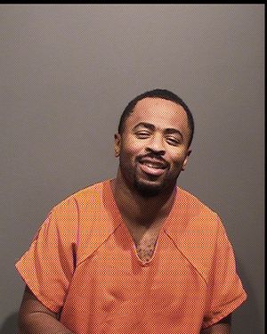 A man wearing an orange prison jumpsuit smiles at the camera against a gray background.