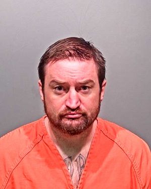 A man with a beard and short hair is shown in a mugshot wearing an orange jumpsuit against a gray background.