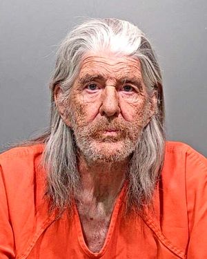 A man with long, gray hair and facial stubble is wearing an orange jumpsuit and looking directly at the camera.