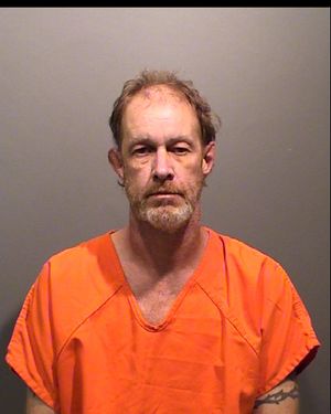 A middle-aged man with a beard is wearing an orange prison jumpsuit and looking directly at the camera against a neutral background.