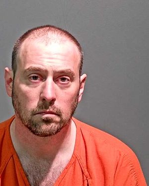 A man with a shaved head and beard is shown in a prison jumpsuit, looking directly at the camera.