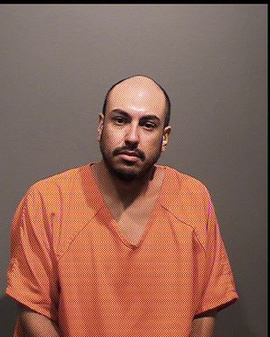A man wearing an orange jail jumpsuit is facing forward, appearing serious, against a plain background.