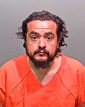 A man with dark hair and a beard is shown wearing an orange prison jumpsuit against a plain gray background.