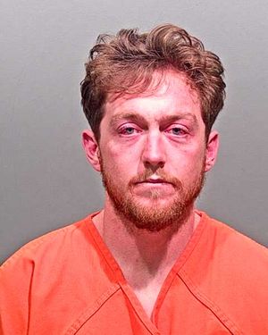 A man with a beard and disheveled hair is shown in an orange jail uniform against a plain background.