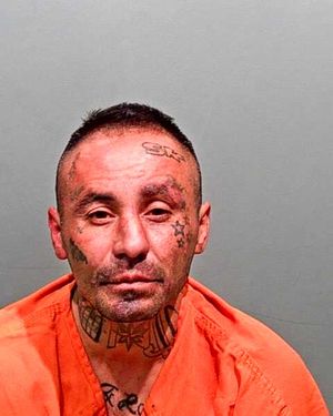 A man with extensive facial tattoos is shown wearing an orange jumpsuit against a plain background.