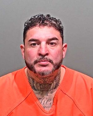 A man with short, curly hair and facial tattoos wearing an orange prison jumpsuit.