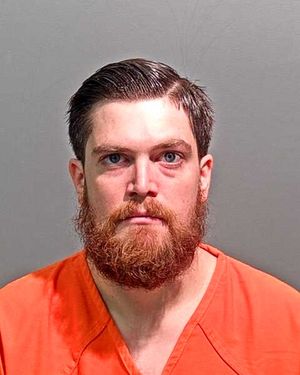 A man with a full beard and short hair is wearing an orange jumpsuit and looking directly at the camera.