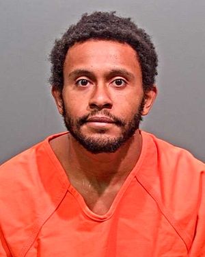 A man with short curly hair and a beard is shown in a mugshot wearing an orange prison jumpsuit.