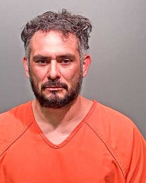 A man with a beard and tousled hair is wearing an orange prison jumpsuit and looking directly at the camera.