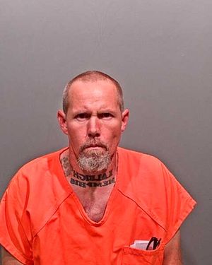 A man in an orange jail uniform with visible tattoos and a serious expression is shown against a gray background.