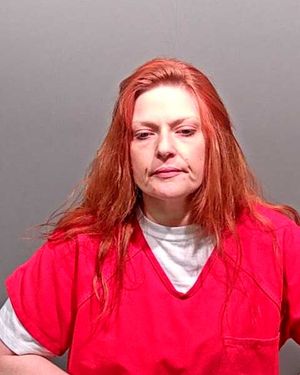 A woman with long red hair is wearing a red prison jumpsuit and appears to be in a mugshot setting.