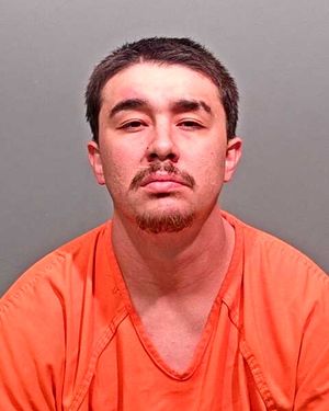 A young man with short hair and facial hair is shown in a mugshot, wearing an orange prison jumpsuit.