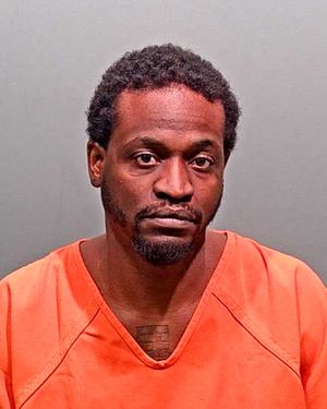 A man wearing an orange jumpsuit is posed for a mugshot against a gray background.