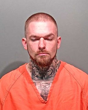 A man with extensive tattoos on his neck and face is wearing an orange inmate jumpsuit and looking downwards.