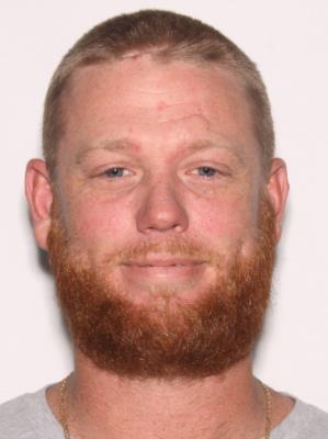 A man with short hair and a full beard smiles for the camera against a plain background.