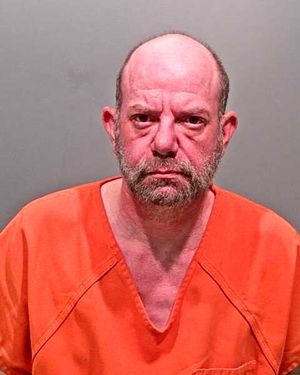A man with a shaved head and a beard is shown wearing an orange jail uniform, looking directly at the camera.