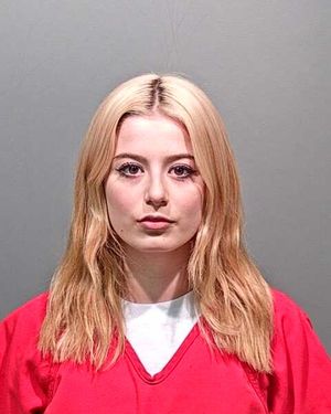 A young woman with long, blonde hair is wearing a red prison jumpsuit in a mugshot setting.