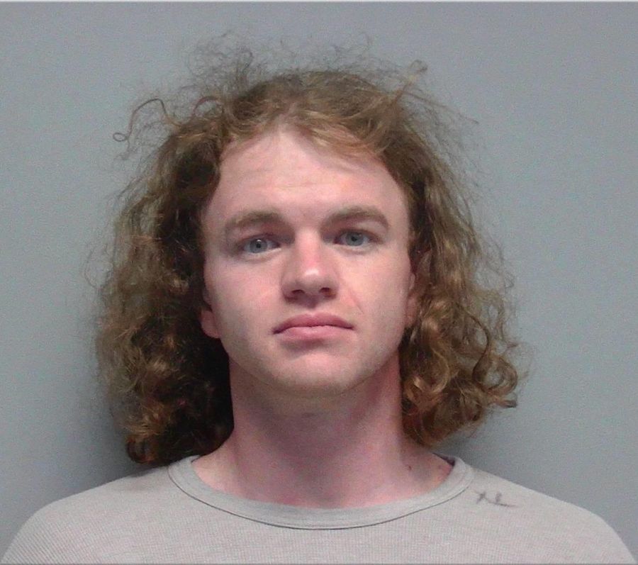 Mugshot-style booking photo of a young man with long curly light-brown hair, wearing a light gray shirt, facing the camera against a plain gray background.