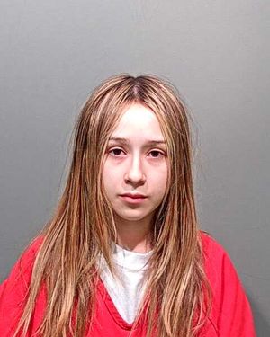 A young person with long hair is shown in a mugshot wearing a red prison uniform against a gray background.