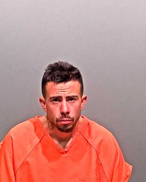 A man wearing an orange prison jumpsuit is seated against a gray background.