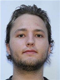 A young man with shoulder-length, light brown hair and a light beard is facing the camera against a plain background.