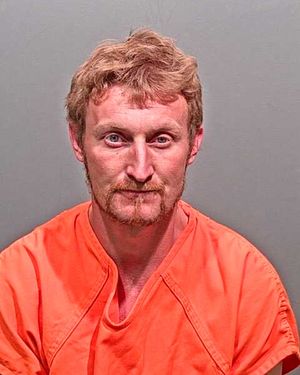 A man with reddish-brown hair and a beard is shown wearing an orange prison jumpsuit against a gray background.